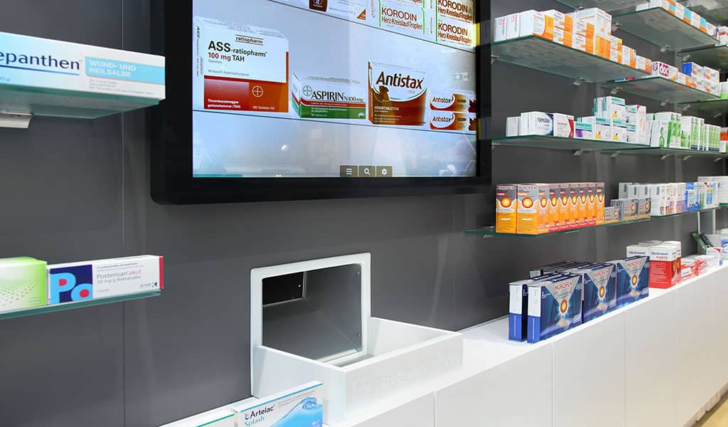 Ausgabe eines Lagerautomat in der Apotheke zwischen mehreren Regalen und einem Werbebildschirm - vitasco GmbH